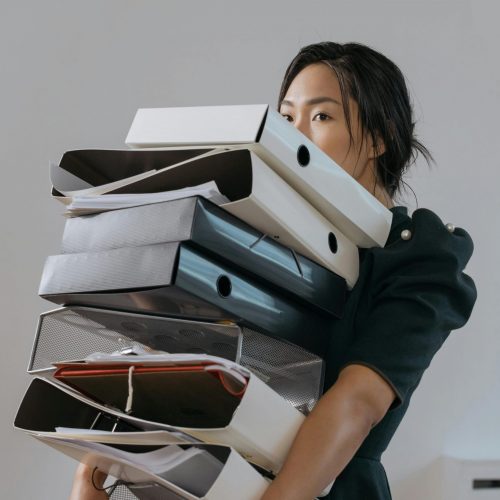 Woman carrying a large stack of file folders indoors, expressing stress and workload.